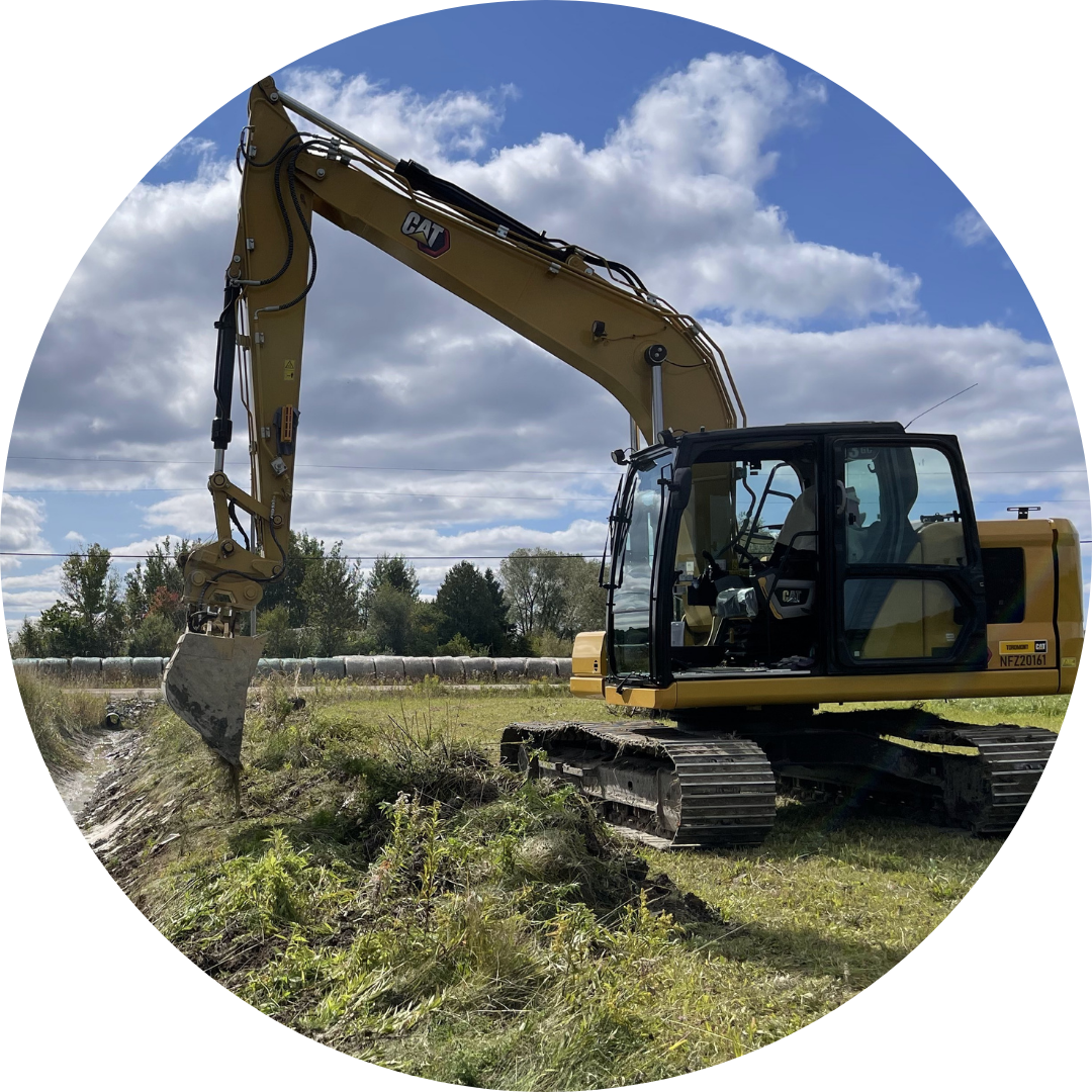 an excavator owned by Proficient Excavating of Sturgeon Falls, Ontario sits in a large field beside a machine-dug ditch